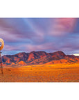Desert landscape with a windmill and mountains under a colorful sky.