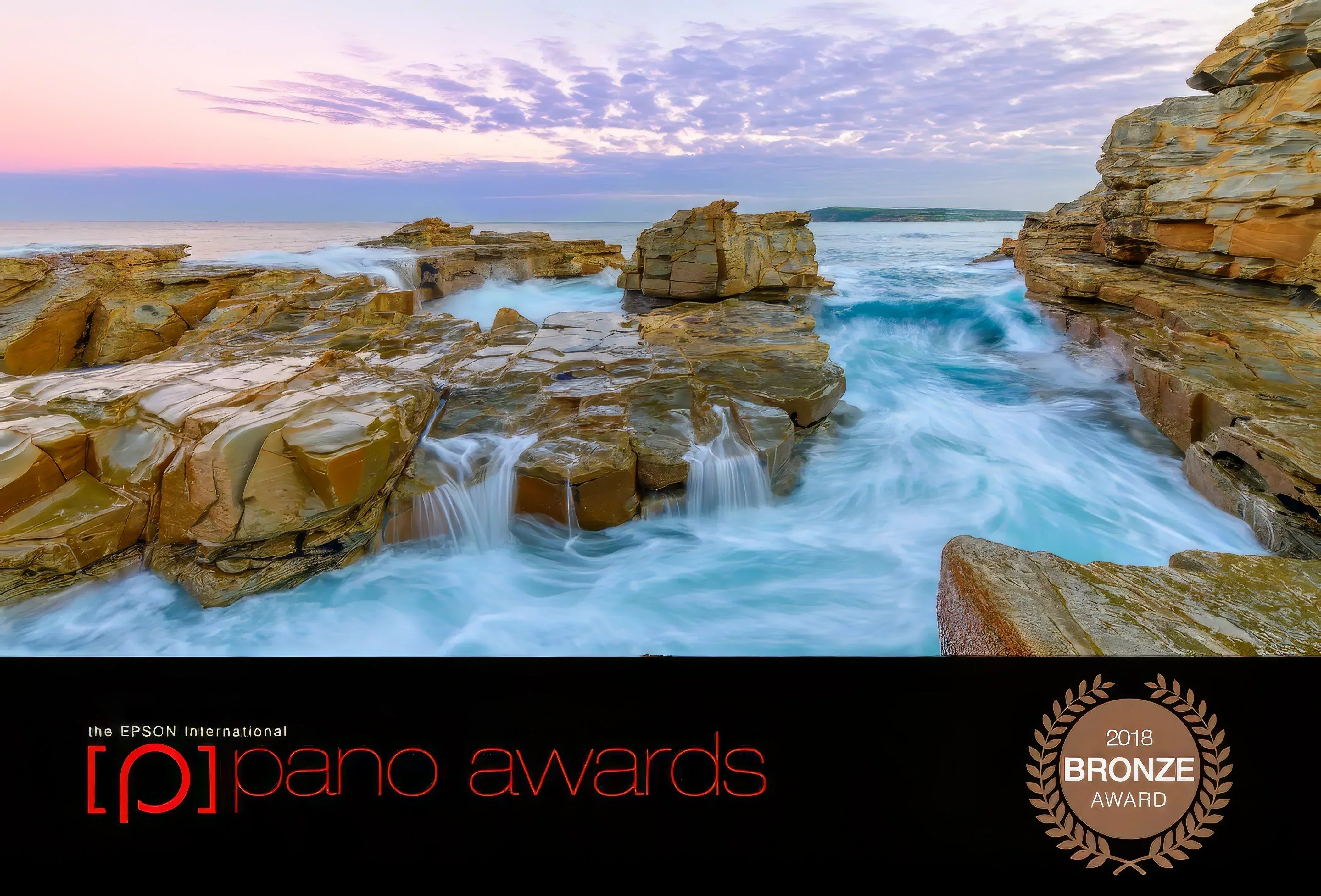Award-winning coastal seascape featuring rugged sandstone rocks and soft flowing waves at sunset, recipient of the 2018 Bronze Award from the Epson International Pano Awards