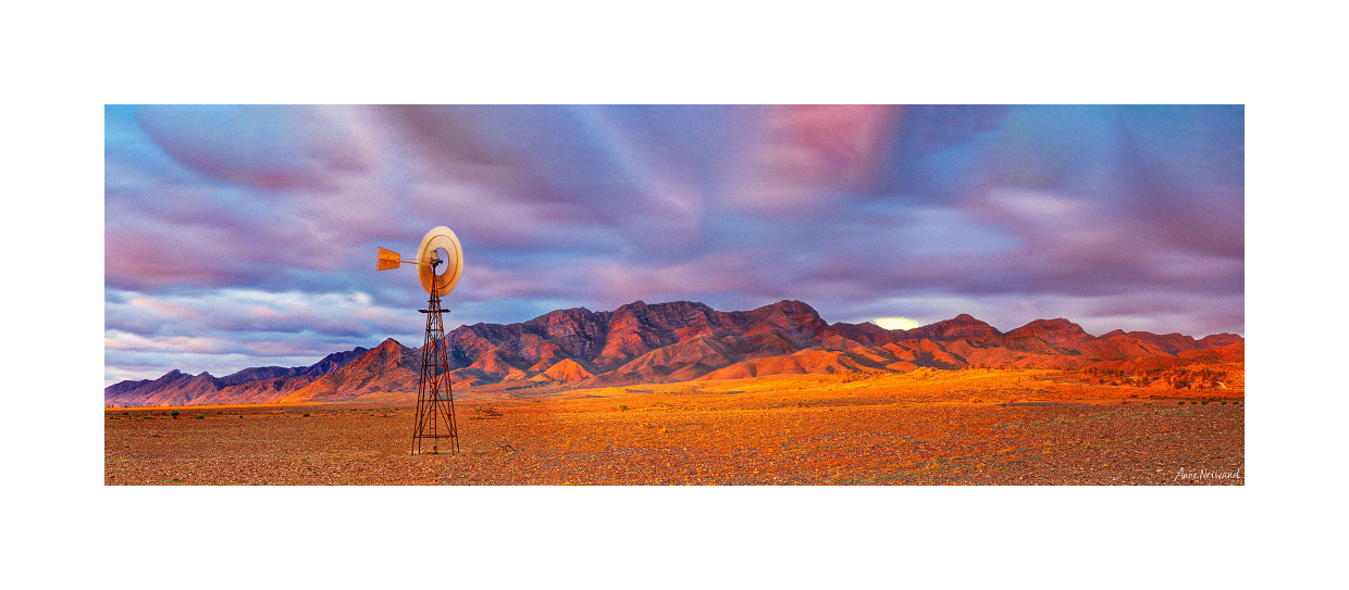 Desert landscape with a windmill and mountains under a colorful sky.